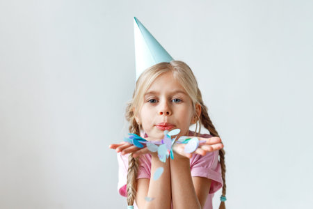 Indoor shot of pretty small kid with long pigtails, wears cone hat on head, blows colourful sheet of...