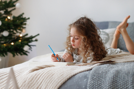 Indoor shot of attentive pretty small girl writes letter to Santa Claus before Christmas, thinks what present she expects, wants all dreams and wishes come true, lies on comfortable bed indoor.の写真素材
