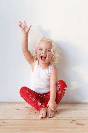 Photo of joyful blonde small kid sits on wooden floor, dressed casually, raises hand, isolated over white background. Cute lovely girl with cheerful expression. Children and emotions concept.の写真素材