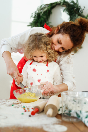 Affectionate mother helps her dughter to whisk eggs in bowl, stand together on kitchen, bake delicious pie for all family. Mum teaches small female child to cook. Culinary preparation for Christmasの写真素材