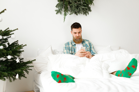 Restful handsome male with long beard and mustache has rest at comfortable bed, surfes social networks on smart phone, spends winter holidays at home, enjoys domestic atmosphere and wireless wifi.の写真素材