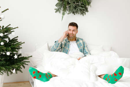 Indoor shot of positive attractive bearded male dressed in casual checkered shirt and green socks, has phone conversation, shares with friend how he spent New Year night, admires decorated firtree.の写真素材