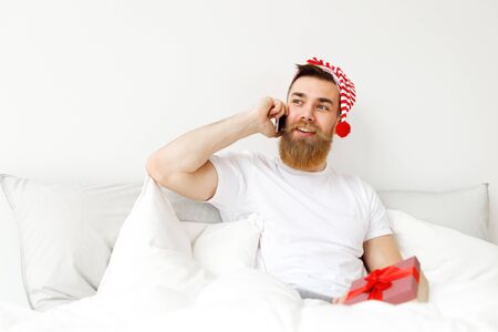 Indoor shot of attractive man with thick long beard and mustache speaks over mobile phone, sits in comfortable bed, prepares wrapped present box for his girlfriend. Holidays, people, technologyの写真素材