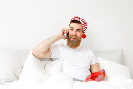 Happy young bearded guy in festive hat has telephone conversation with someone, holds wrapped present box, covered with white warm blanket in bed, enjoys good morning and domestic atmosphereの写真素材