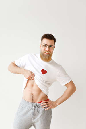 Handsome muscular man raises his white t shirt, demonstrates naked body, poses against white studio background. Positive bearded male model in spectacles shows perfect masculine body. Vertical shotの写真素材
