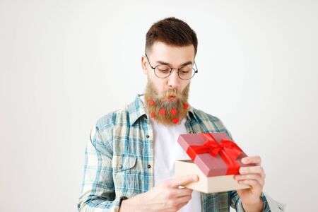 Surprsied handsome man with thick beard and mustache looks with intriguing expression as opens wrapped gift box, being curious what is there, wears round spectacles and checkered shirt, isolatedの写真素材