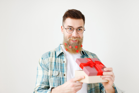 Fashionable bearded hipster guy in eyewear, opens wrapped present, recieves surprise from wife, being in good festive mood, isolated over white background. People, surprisment and presents conceptの写真素材