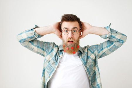 Stupefied surprised man with long beard dressed in fashionable clothes, keeps hand on head, stares through spectacles, being amazed to hear shocking news, isolated over white studio backgroundの写真素材