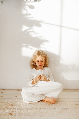 Restful adorable small kid dressed in long white dress, plays with Christmas toy deer, sits crossed legs on wooden floor, isolated over white studio background. Happy child plays with favourite toy.の写真素材