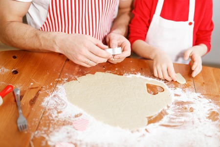 Cropped shot of unrecognizable hard working father and small daughter sit together at wooden table, make forms from dough, prepared puff pastry, surrounded with flour. Baking and cooking concept.の写真素材