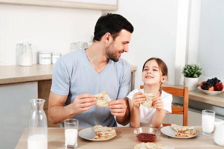 Affectionate pleasant looking father and his little daughter eat tasty morning meal at kitchen, communicate with each other.の写真素材