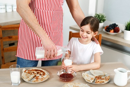 Cropped image of careful father wears apron, pours fresh milk in glass for his daughter, bakes delicious pancakes, prepares breakfast for child, being together at kitchen. Family, food, children.の写真素材
