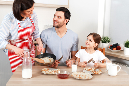 Photo of hard working wife, husband and their daughter sit together at kitchen table, going to eat delicious pancakes, enjoy togetherness.の写真素材