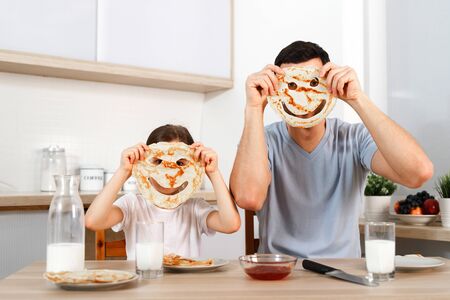 Funny pretty daughter and father make faces with pancakes, make fun together at kitchen during weekends, enjoy delicious flannel cake, foolish.の写真素材