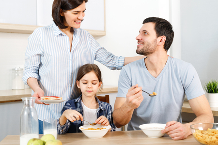 Three family members have delicious healthy breakfast at kitchen, eat cornflakes with milk, enjoy togetherness and domestic atmosphere.の写真素材