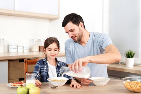 Young handsome father pours milk in bowl with flakes, prepares breakfast for small child, sit together at kitchen, have healthy food, enjoy togetherness. Friendly family being at home, eat.の写真素材