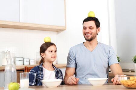 Funny young dad and daughter sit next to each other, eat delicious cornflakes, keep apples on head, demonstrate that they eat only healthy food, have good relationships and understanding.の写真素材