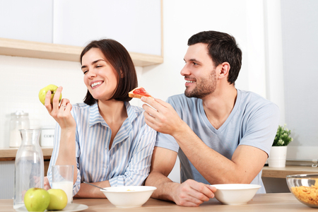Happy couple spend free time or weekend together at kitchen, glad husband suggests wife to eat snack, she refuses as eats green apple, have breakfast. Family, cooking and relations concept.の写真素材