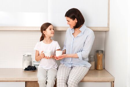 Shot of cheerful mother and daughter sit together at kitchen table, drink hot tea in morning, have pleasant friendly talk between each other.の写真素材