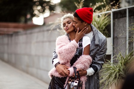 Loving mulatto couple embrace each other while have walk outdoor, kiss. Dark skinned stylish male in trendy red hat hugs his girlfriend with specific appearance. People, ethicity, relationship concept.の写真素材