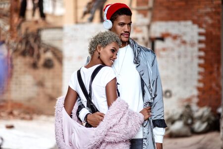 Mixed race confident stylish male wears headphones, embraces his mulatto girlfriend, pose together against brick wall with thoughtful expression, enjoy spare time. African American couple in love.の写真素材