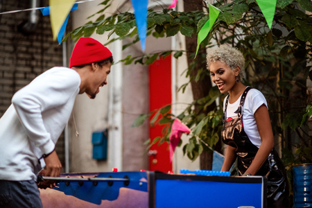 Two mixed race female and male friends play table football outdoor, entertain themselves. Positive stylish mulatto people play outdoor game together. People, recreation and entertainment concept.の写真素材