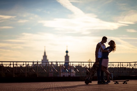 Outdoor shot of affectionate young couple pose against beautiful city view and sky, have rest after skateboading, demonstrate true love and perfect relationships. Talented skateboarders togehter.の写真素材