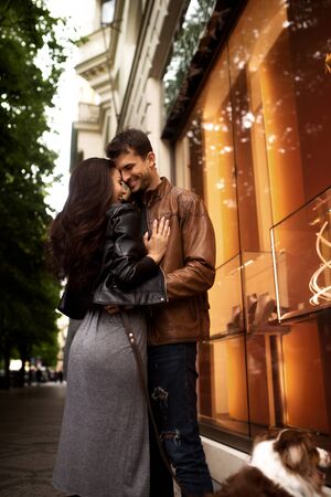 Vertical portrait of happy lovely couple stand close to each other, have walk with dog, enjoy togetherness and good rest, breath fresh air. Handsome man embraces his elegant brunette girlfriend.の写真素材