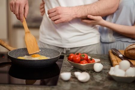 Unrecognizable female and male fry together eggs and make tomatoes salad, stand on kitchen, prepare breakfast involved in cooking. Cropped shot of friendly family couple make delicious dinner.の写真素材