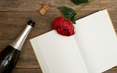 An open book with blank pages, a red rose with green leaves, an open bottle of sparkling wine on a wooden background. Text space. Celebrations, Valentine's Day. Minimal style. Top view. Flat lay.の写真素材
