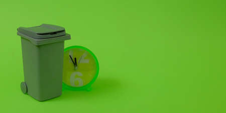 Trash can and a clock showing 5 minutes before 12 on a bright green background. Text space. Environmental protection concept. Minimal style.の写真素材