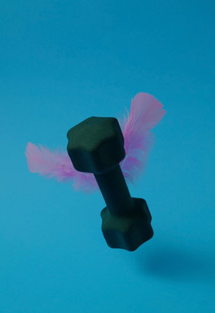 Dark green dumbbell with soft light pink feathers in the shape of wings floats on a bright blue background. Text space. Exercise is easy, concept. Minimal style.の写真素材