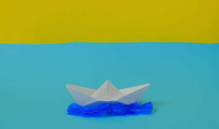 Boat made of white paper on blue feathers on a bright blue and yellow background. Text space. Holiday time, travel, adventure, float away, creative, concept. Minimal style.の写真素材