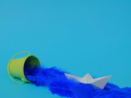 Spilled blue feathers from a green bucket and a boat made of white paper on a bright blue background.. Text space. Time for travel, vacation, creative concept. Minimal style.の写真素材
