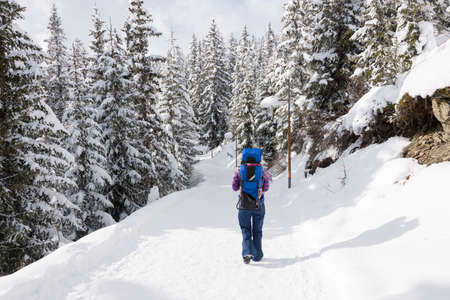 Tourist mother in ski gear walking in the snow in a forest with snow on the trees with her baby child on her back in a baby carrier backpack in a ski resort area in winter.の写真素材