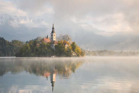 Church on the small island in Lake Bled and Bled Castle in the first sunlight of sunrise on a beautiful foggy morning in autumn with reflection in the calm water in Slovenia.の写真素材