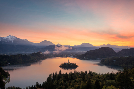 View over Bled lake at sunrise with the island with the church, mountains with snow in the background and reflection of the colorful sky in the lake in a landscape in Slovenia.の写真素材