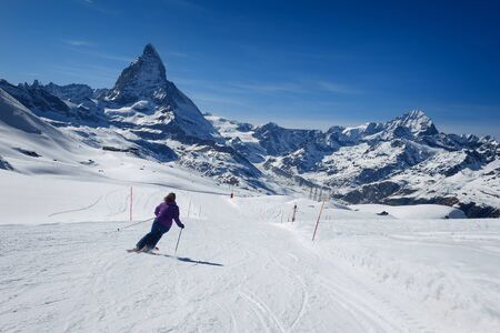 A female skier skiing without a helmet alone on the ski slope towards majestic Matterhorn mountain on a beautiful sunny day with a thick layer of snow in ski resort Zermatt, canton of Valais in Switzerland.の写真素材