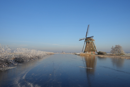 A typical winter landscape in the Netherlands with a windmill and hoar frosted reeds along the ice of a frozen river on a beautiful clear sky day in the dutch countryside near Kinderdijk.の写真素材