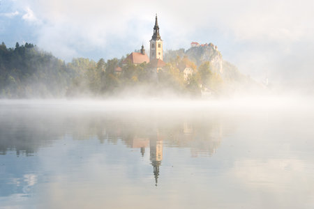 A layer of fog over the still water of Bled Lake in front of the catholic church of Mary the Queen...