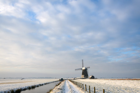 Minimalistic view on the dutch countryside in winter with a dirt road with a fence and a ditch towards a traditional windmill, covered in snow under a nice sky in the early morning light near Kinderdyk in the Netherlands.の写真素材