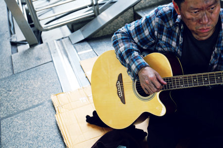 Homeless man sitting and playing acoustic guitar on walking street in the capital city.