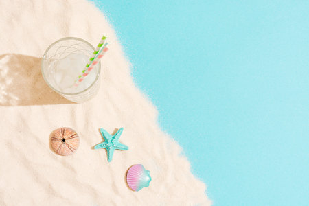 Summer concept composed of tall glass with ice and straws and of sea urchin, starfish and a shell laid out on sand and blue background.の写真素材