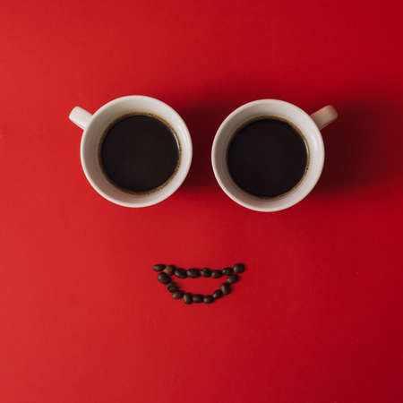 Happy face composed of two coffee cups and beans laid out on red background. Minimal flat lay.の写真素材
