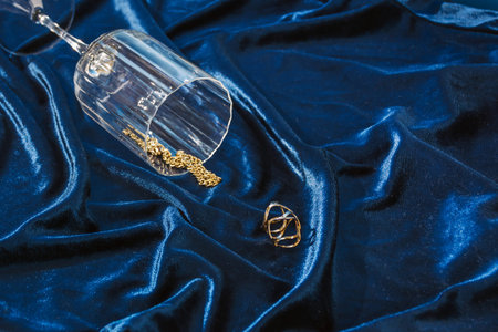 Golden ring and chain spilling from tall glass of drink on blue velvet background from angle.