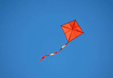 A vibrant red kite soars high in a clear blue sky its long tail streaming behind it.の素材