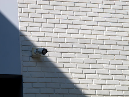 A surveillance camera is installed on a white brick wall, casting a prominent shadow. This image highlights security technology and architectural design.の写真素材