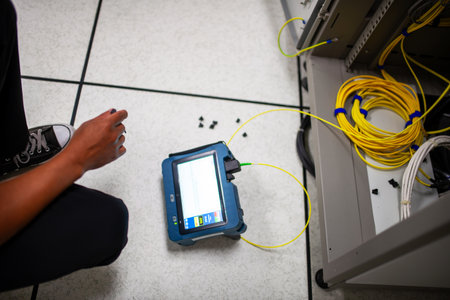 A technician kneels beside network equipment using a tablet for testing and diagnostics. Yellow cables surround the workspace, highlighting technology in action.の写真素材