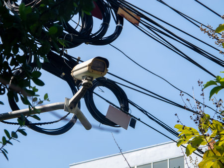 A surveillance camera mounted overhead amid tangled cables in an urban setting. The scene captures modern security measures integrated into city infrastructure.の写真素材