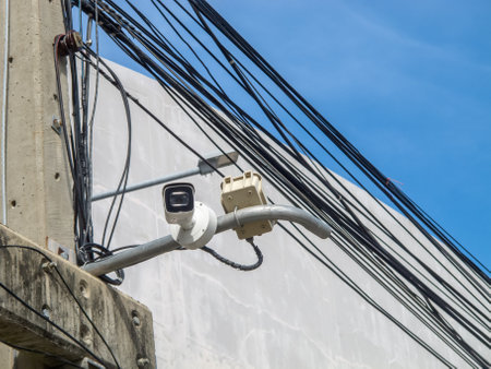 A security camera is mounted on a utility pole, surrounded by power lines and wires, set against a bright blue sky, showcasing urban infrastructure and technology.の写真素材